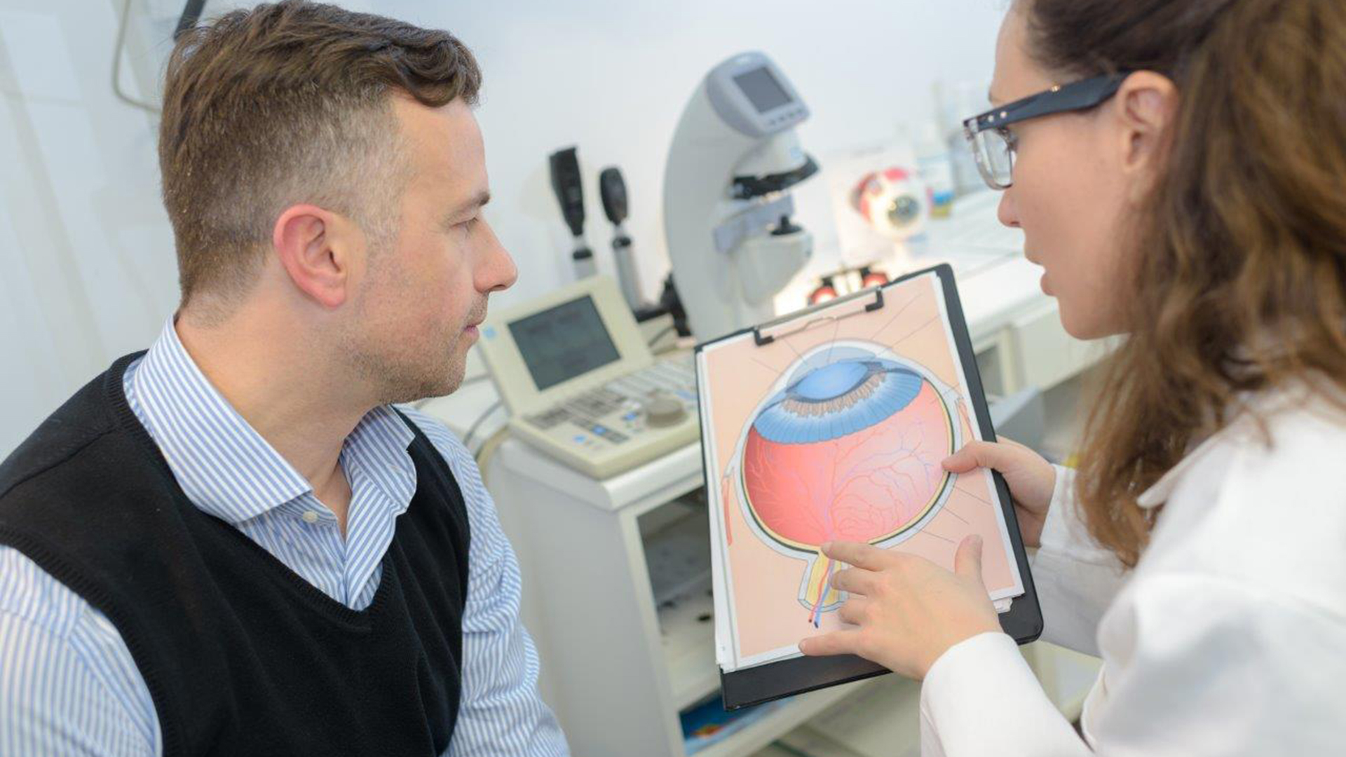 Optometristin erklärt anhand eines Bildes vom Auge einem Patienten den Befund