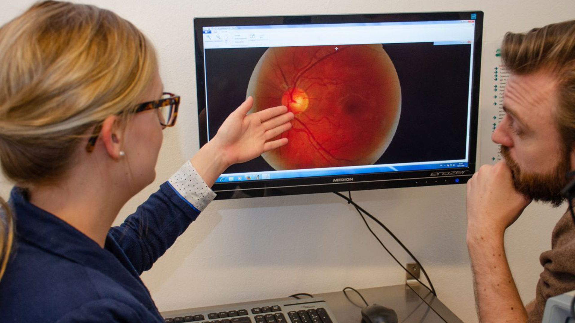 Optometristin zeigt einem Patienten Aufnahmne des Augenhintergrundes am Bildschirm